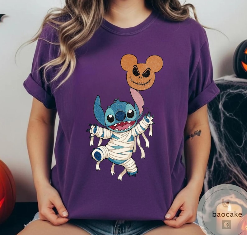 May include: Purple t-shirt featuring a Halloween-themed graphic. The design includes a blue cartoon character wrapped in bandages, holding a jack-o'-lantern balloon. The jack-o'-lantern is brown with a carved face. The shirt has short sleeves.