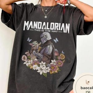 Puede incluir: Camiseta gris oscuro con el texto blanco "MANDALORIAN - THIS IS THE WAY". El dise&ntilde;o muestra un personaje mandaloriano y Grogu, rodeados de flores y mariposas. La camiseta es de corte holgado.