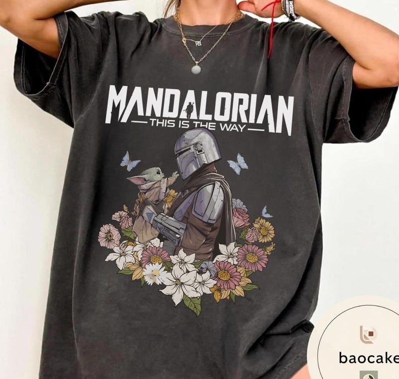 Puede incluir: Camiseta gris oscuro con el texto blanco "MANDALORIAN - THIS IS THE WAY". El dise&ntilde;o muestra un personaje mandaloriano y Grogu, rodeados de flores y mariposas. La camiseta es de corte holgado.