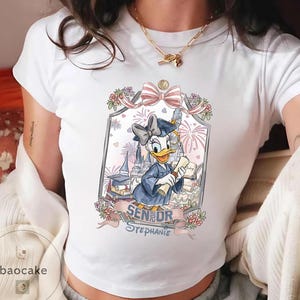 May include: White t-shirt featuring a graduation-themed graphic of Daisy Duck in a cap and gown, holding a diploma. The design includes a castle, fireworks, and the text "Senior Stephanie."