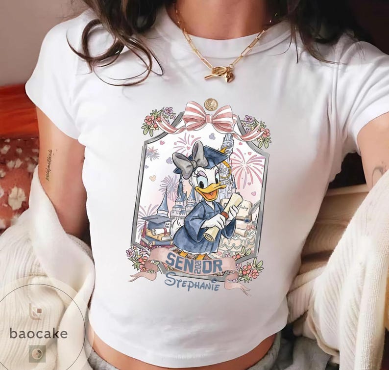 May include: White t-shirt featuring a graduation-themed graphic of Daisy Duck in a cap and gown, holding a diploma. The design includes a castle, fireworks, and the text "Senior Stephanie."