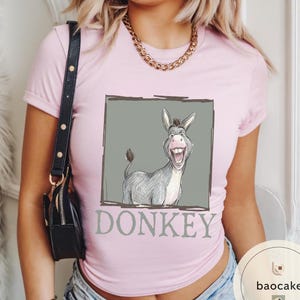 May include: A light pink t-shirt featuring a cartoon donkey illustration inside a gray rectangle. The donkey is gray and white with a large smile. The word "DONKEY" is printed below the image in gray.