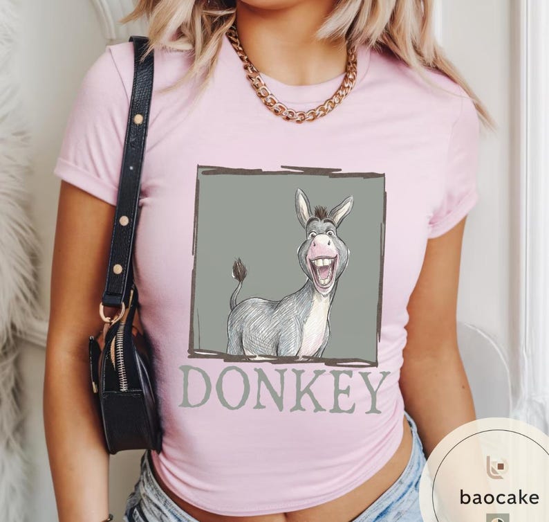 May include: A light pink t-shirt featuring a cartoon donkey illustration inside a gray rectangle. The donkey is gray and white with a large smile. The word "DONKEY" is printed below the image in gray.