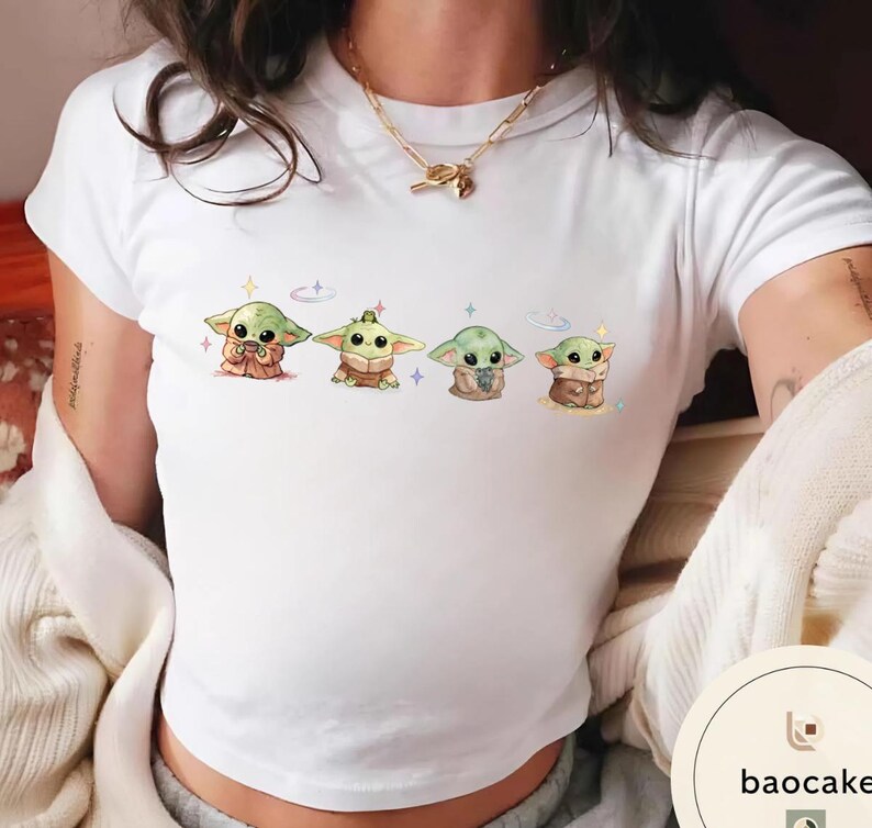 May include: White t-shirt featuring a colorful illustration of four Grogu characters from the Star Wars series. Each character is depicted in a different pose, surrounded by stars and sparkles. The design is centered on the front of the shirt.