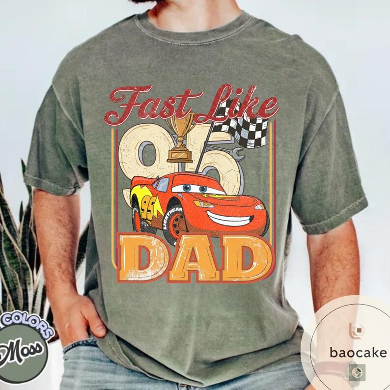 May include: Olive green t-shirt featuring a graphic of a red race car with the text "Fast Like Dad". The design includes a trophy, checkered flag, and the number 95. The shirt is a comfortable, casual wear.