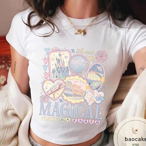 May include: White t-shirt with a colourful graphic featuring theme park icons, including a castle, a globe, and a monorail. The text "MAGICAL PLACE ON EARTH" is displayed in a playful font, with pastel colours.