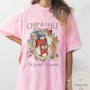 May include: Pink t-shirt featuring Chip and Dale in wizard costumes, surrounded by a floral wreath. The text "Chip & Dale" is above the image, and "Wizard Wonders" is below. The shirt has white trim on the sleeves.