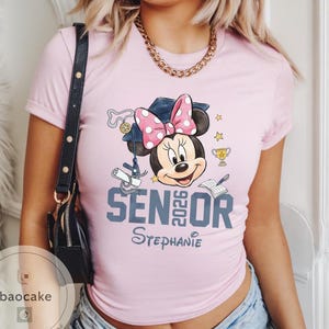 May include: A light pink t-shirt featuring a Minnie Mouse graphic wearing a graduation cap with a pink polka dot bow. The shirt says "SENIOR 2026" and "Stephanie" in a script font.