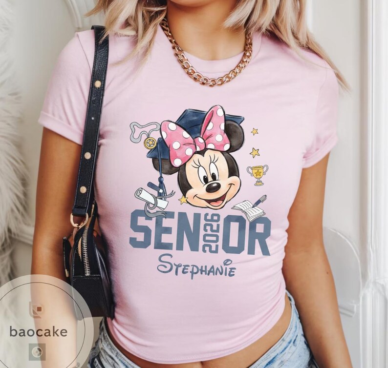 May include: A light pink t-shirt featuring a Minnie Mouse graphic wearing a graduation cap with a pink polka dot bow. The shirt says "SENIOR 2026" and "Stephanie" in a script font.