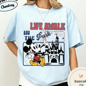 May include: Pale blue t-shirt with a graphic of Mickey Mouse and the text "LIFE A WALK IN THE Park". The design includes a castle, fireworks, and a blue border. The shirt is made of a soft material.