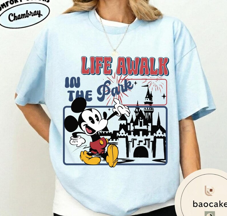 May include: Pale blue t-shirt with a graphic of Mickey Mouse and the text "LIFE A WALK IN THE Park". The design includes a castle, fireworks, and a blue border. The shirt is made of a soft material.