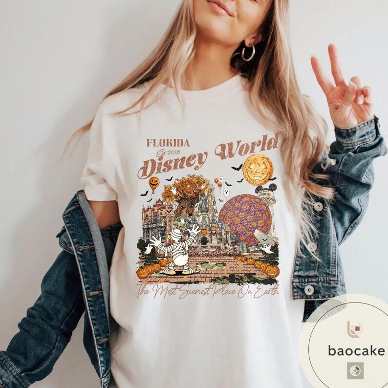Op de afbeelding: Cr&egrave;mekleurig T-shirt met een Halloween-thema Disney World-afbeelding. Het ontwerp bevat een mummie, pompoenen en een spookkasteel. De tekst luidt "Florida Disney World" en "The Most Scariest Place On Earth."