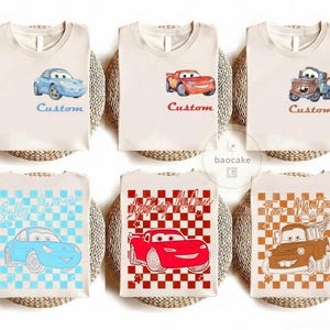 May include: Six beige t-shirts with cartoon car graphics. The shirts feature characters like Lightning McQueen, Sally Carrera, and Tow Mater. Each shirt has the word "Custom" or a character's name. The designs are in blue, red, and brown.