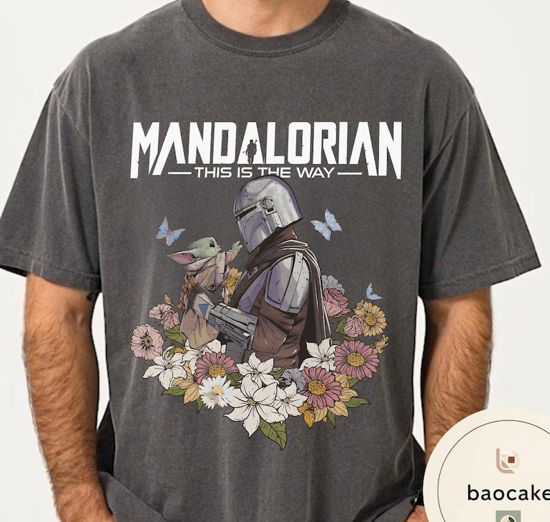 Puede incluir: Camiseta gris oscuro con un dise&ntilde;o de The Mandalorian y Grogu rodeados de flores y mariposas. El texto "MANDALORIAN - THIS IS THE WAY" est&aacute; impreso en blanco. El dise&ntilde;o incluye una ilustraci&oacute;n detallada de los personajes.