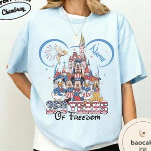 Peut inclure: T-shirt bleu clair avec un motif Disney repr&eacute;sentant des personnages devant un ch&acirc;teau, avec le texte "250 Years of Freedom". Le motif comprend Mickey Mouse, Donald Duck et d'autres personnages. Le mot "Name" est &eacute;galement visible.