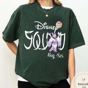 Puede incluir: Camiseta verde oscuro con la frase "Disney Squad" en blanco, con un dibujo de Daisy Duck con un kimono morado sosteniendo un globo arco&iacute;ris. Las palabras "Big Sis" est&aacute;n debajo.