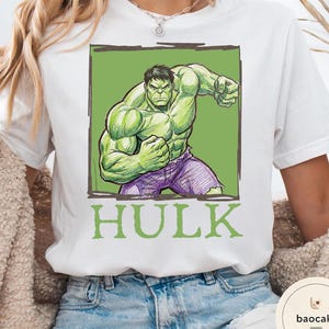 May include: White t-shirt featuring a green and purple illustration of the Hulk in a fighting pose, set against a green background. The word "HULK" is printed in green below the image.