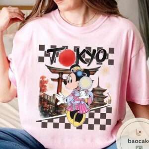 Puede incluir: Camiseta rosa claro con un dise&ntilde;o de Minnie Mouse con kimono, sosteniendo un abanico, y la palabra "TOKYO". El dise&ntilde;o incluye un templo japon&eacute;s y un patr&oacute;n de tablero de ajedrez en blanco y negro.