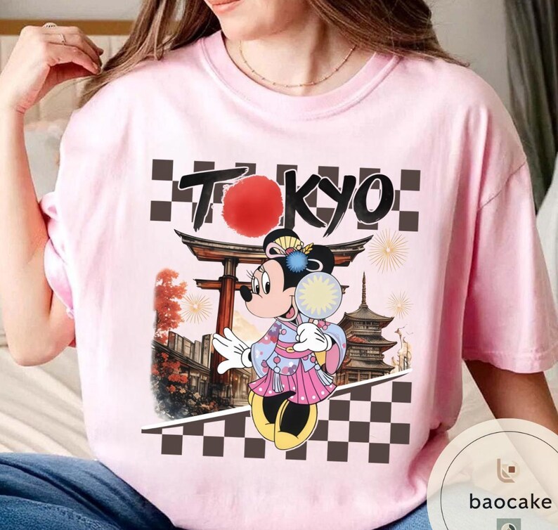 Puede incluir: Camiseta rosa claro con un dise&ntilde;o de Minnie Mouse con kimono, sosteniendo un abanico, y la palabra "TOKYO". El dise&ntilde;o incluye un templo japon&eacute;s y un patr&oacute;n de tablero de ajedrez en blanco y negro.