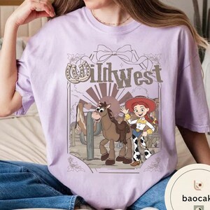 Pu&ograve; includere: T-shirt lilla chiaro con un design del Far West. La grafica include un cavallo dei cartoni animati, una cowgirl e la scritta "Wild West". Il design &egrave; ambientato in un deserto con cactus.