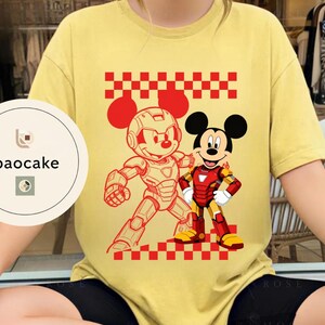 May include: A yellow t-shirt featuring a red and white checkered pattern and a graphic of Mickey Mouse in an Iron Man-inspired suit and another in a robotic suit. The shirt has short sleeves and a round neckline. The brand logo "baocake" is visible on the left side.