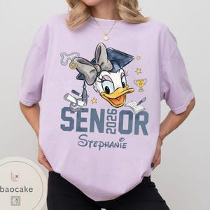 May include: A light purple t-shirt featuring a Daisy Duck graduation design. The design includes a graduation cap, bow, diploma, trophy, stars, and the text "SENIOR 2026 Stephanie". The shirt is a comfortable, casual style.