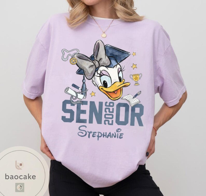 May include: A light purple t-shirt featuring a Daisy Duck graduation design. The design includes a graduation cap, bow, diploma, trophy, stars, and the text "SENIOR 2026 Stephanie". The shirt is a comfortable, casual style.