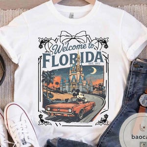 May include: White t-shirt featuring a graphic of Mickey Mouse driving a red convertible on a road towards a castle. The text "Welcome to FLORIDA EST. 1971" is displayed above the graphic in a decorative frame.