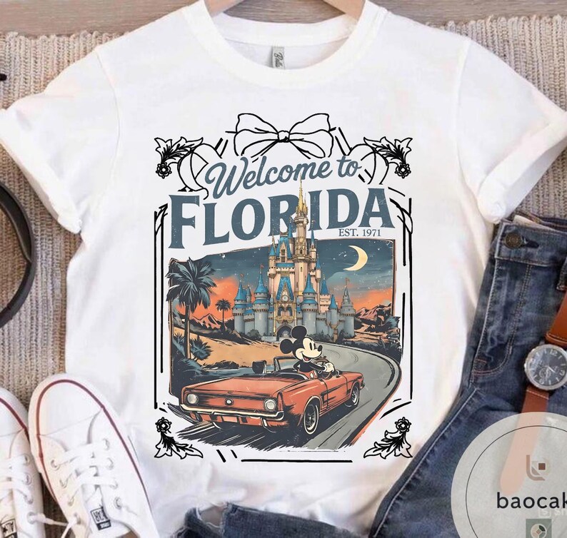 May include: White t-shirt featuring a graphic of Mickey Mouse driving a red convertible on a road towards a castle. The text "Welcome to FLORIDA EST. 1971" is displayed above the graphic in a decorative frame.