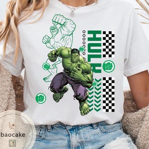 May include: White t-shirt featuring a graphic of the Hulk in action, with green and purple tones. The word "HULK" is vertically printed in green, along with a green fist symbol and a checkered pattern. The shirt has a crew neck.