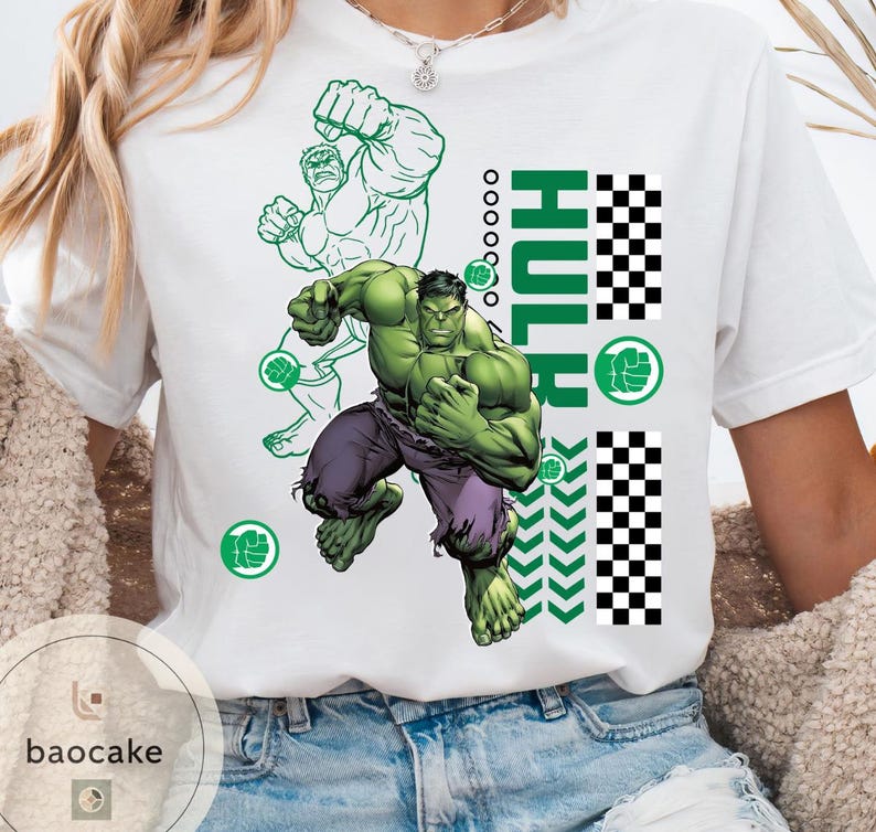 May include: White t-shirt featuring a graphic of the Hulk in action, with green and purple tones. The word "HULK" is vertically printed in green, along with a green fist symbol and a checkered pattern. The shirt has a crew neck.