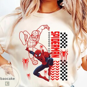 May include: Cream-colored t-shirt featuring a graphic of Spider-Man in action, with red and blue colors. The design includes the word "SPIDERMAN" in red, along with spider symbols and a checkered pattern. The shirt has a crew neck.