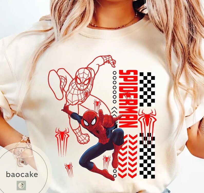 May include: Cream-colored t-shirt featuring a graphic of Spider-Man in action, with red and blue colors. The design includes the word "SPIDERMAN" in red, along with spider symbols and a checkered pattern. The shirt has a crew neck.