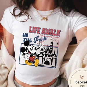 May include: White t-shirt with a graphic of Mickey Mouse gesturing towards a castle, and the text "LIFE A WALK IN THE Park". The design uses red, blue, and yellow, creating a vibrant look. The shirt is a classic crew neck style, suitable for casual wear.