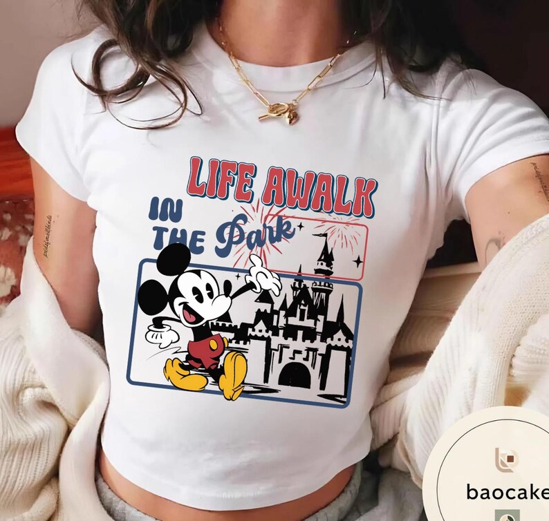 May include: White t-shirt with a graphic of Mickey Mouse gesturing towards a castle, and the text "LIFE A WALK IN THE Park". The design uses red, blue, and yellow, creating a vibrant look. The shirt is a classic crew neck style, suitable for casual wear.