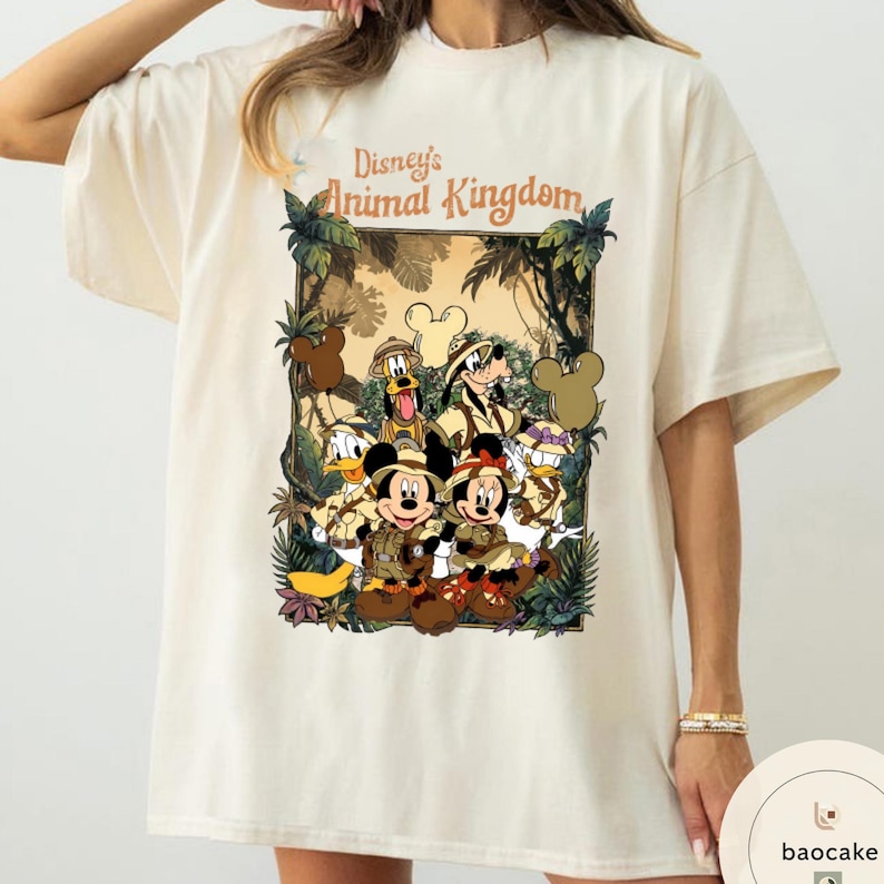 May include: Cream-colored t-shirt featuring a graphic of Disney's Animal Kingdom characters. The design includes Mickey and Minnie Mouse, Donald Duck, Goofy, and other characters in safari attire, surrounded by jungle foliage.