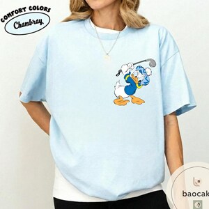May include: Light blue t-shirt with a cartoon graphic of Donald Duck playing golf. The shirt has short sleeves and a crew neck. The words "COMFORT COLORS Chambray" are printed on the upper left side.