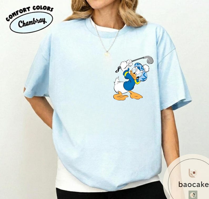 May include: Light blue t-shirt with a cartoon graphic of Donald Duck playing golf. The shirt has short sleeves and a crew neck. The words "COMFORT COLORS Chambray" are printed on the upper left side.