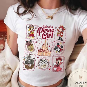 May include: White t-shirt featuring a colorful graphic design with the text "Life of a Pirate Girl." The design includes illustrations of a pirate ship, treasure chest, castle, and Minnie Mouse in pirate attire.
