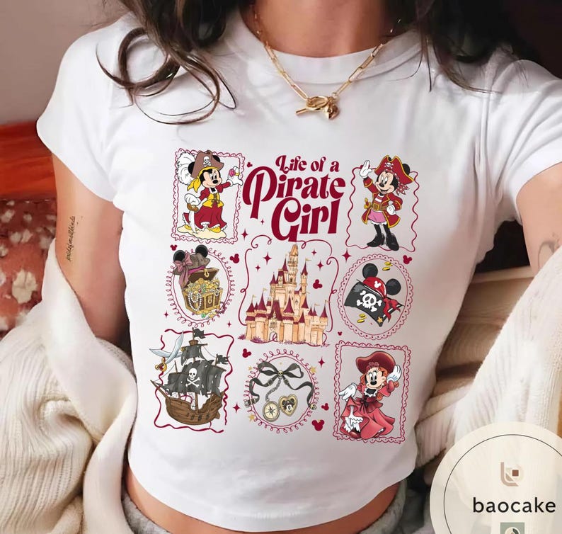 May include: White t-shirt featuring a colorful graphic design with the text "Life of a Pirate Girl." The design includes illustrations of a pirate ship, treasure chest, castle, and Minnie Mouse in pirate attire.