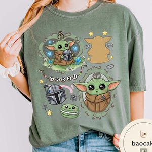 May include: A sage green t-shirt featuring a graphic print of the character Grogu in various poses, a silver helmet, a green macaron, and the text GROGU on a beaded string. The design includes small stars, blue butterflies, and crystals.