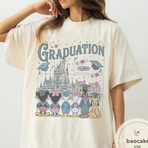 May include: Cream-colored t-shirt featuring a graphic with the word "Graduation" in blue script. The design includes cartoon characters, a castle, graduation caps, and scrolls. The shirt has short sleeves and a crew neck.
