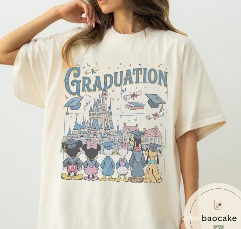 May include: Cream-colored t-shirt featuring a graphic with the word "Graduation" in blue script. The design includes cartoon characters, a castle, graduation caps, and scrolls. The shirt has short sleeves and a crew neck.