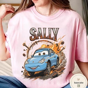 May include: A pale pink t-shirt with a cartoon illustration of a blue car, the name "SALLY" above in a distressed style. The design includes sparks and tools, suggesting a racing theme.