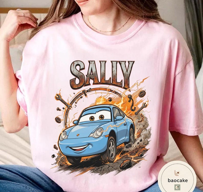 May include: A pale pink t-shirt with a cartoon illustration of a blue car, the name "SALLY" above in a distressed style. The design includes sparks and tools, suggesting a racing theme.