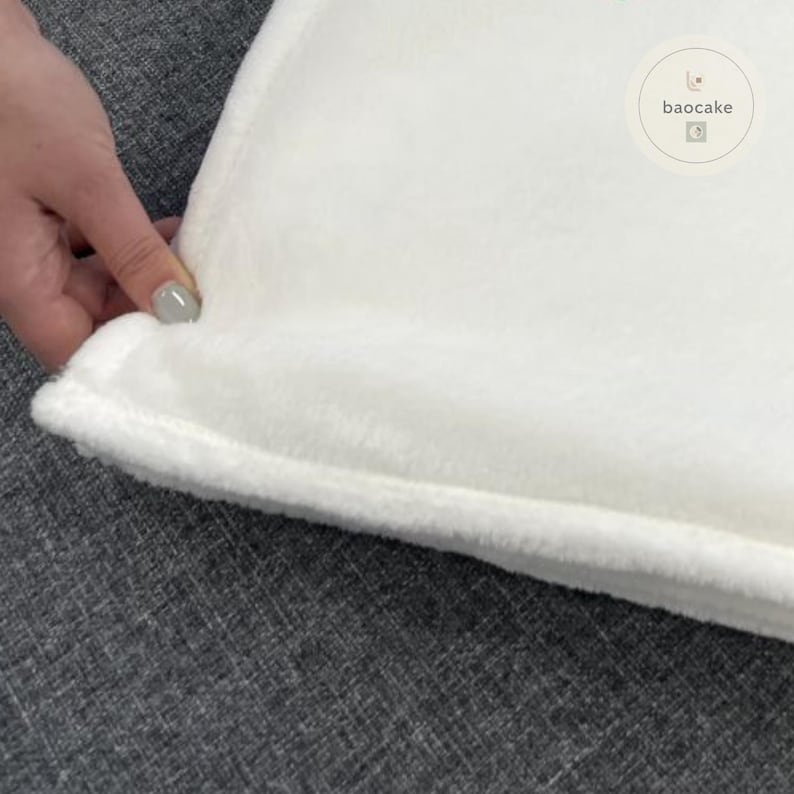 May include: A soft, white plush blanket with a thick, fuzzy texture and finished edges. The fabric has a dense, velvety pile. A circular logo in the top right corner features the text baocake with a small icon above the word.