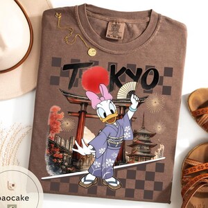 Puede incluir: Camiseta marr&oacute;n con un dise&ntilde;o de Daisy Duck con kimono, sosteniendo un abanico, con la palabra "TOKYO" encima. La camiseta tiene un dise&ntilde;o de tem&aacute;tica japonesa con un sol rojo y una pagoda.