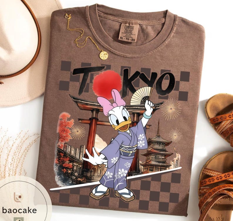 Puede incluir: Camiseta marr&oacute;n con un dise&ntilde;o de Daisy Duck con kimono, sosteniendo un abanico, con la palabra "TOKYO" encima. La camiseta tiene un dise&ntilde;o de tem&aacute;tica japonesa con un sol rojo y una pagoda.
