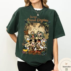 May include: Dark green t-shirt featuring a graphic of Disney's Animal Kingdom characters, including Mickey and Minnie Mouse, Donald Duck, Goofy, and Pluto. The design is framed with jungle foliage and the text "Disney's Animal Kingdom".