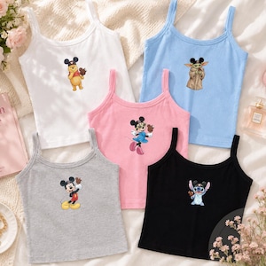 May include: A collection of five sleeveless tank tops in white, light blue, pink, gray, and black. Each top features a small graphic print of a different Disney character wearing Mickey Mouse ears, including Winnie the Pooh, Grogu, Minnie Mouse, Mickey Mouse, and Stitch.